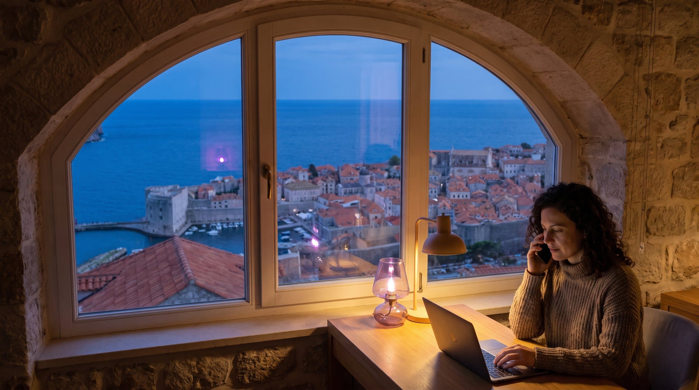 Kamene gradske zidine i crveni krovovi Dubrovnika iznad Jadrana u sumrak, žena telefonira uz osvijetljeni prozor