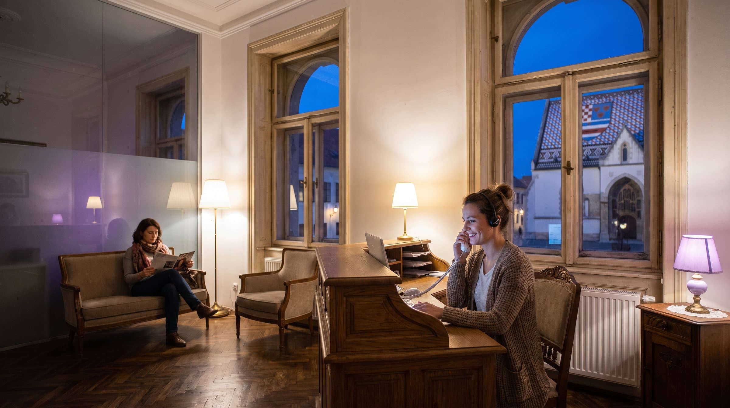 Recepcionarka sa slušalicama javlja se na telefon na recepciji stomatološke ordinacije u zgradi na Gornjem gradu u Zagrebu, kroz prozor se vidi crkva sv. Marka sa šarenim krovom u sumrak